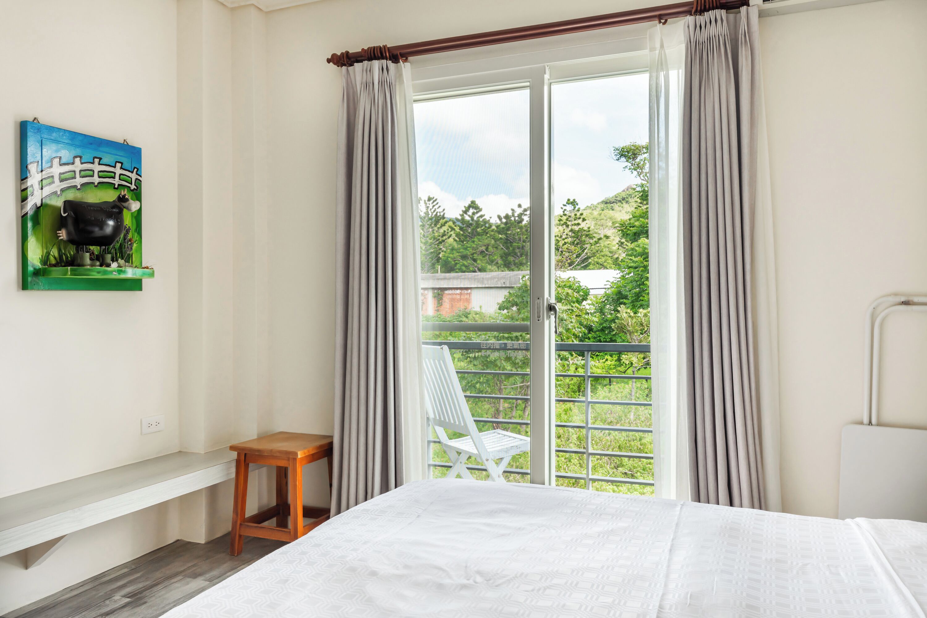 A bedroom with a bed, a wooden nightstand, a chair, a window with curtains, and a painting on the wall.