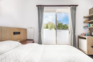 A hotel room with a bed, a wooden headboard, a bedside table, a nightstand, a desk with a chair, a small refrigerator, and a window with curtains.
