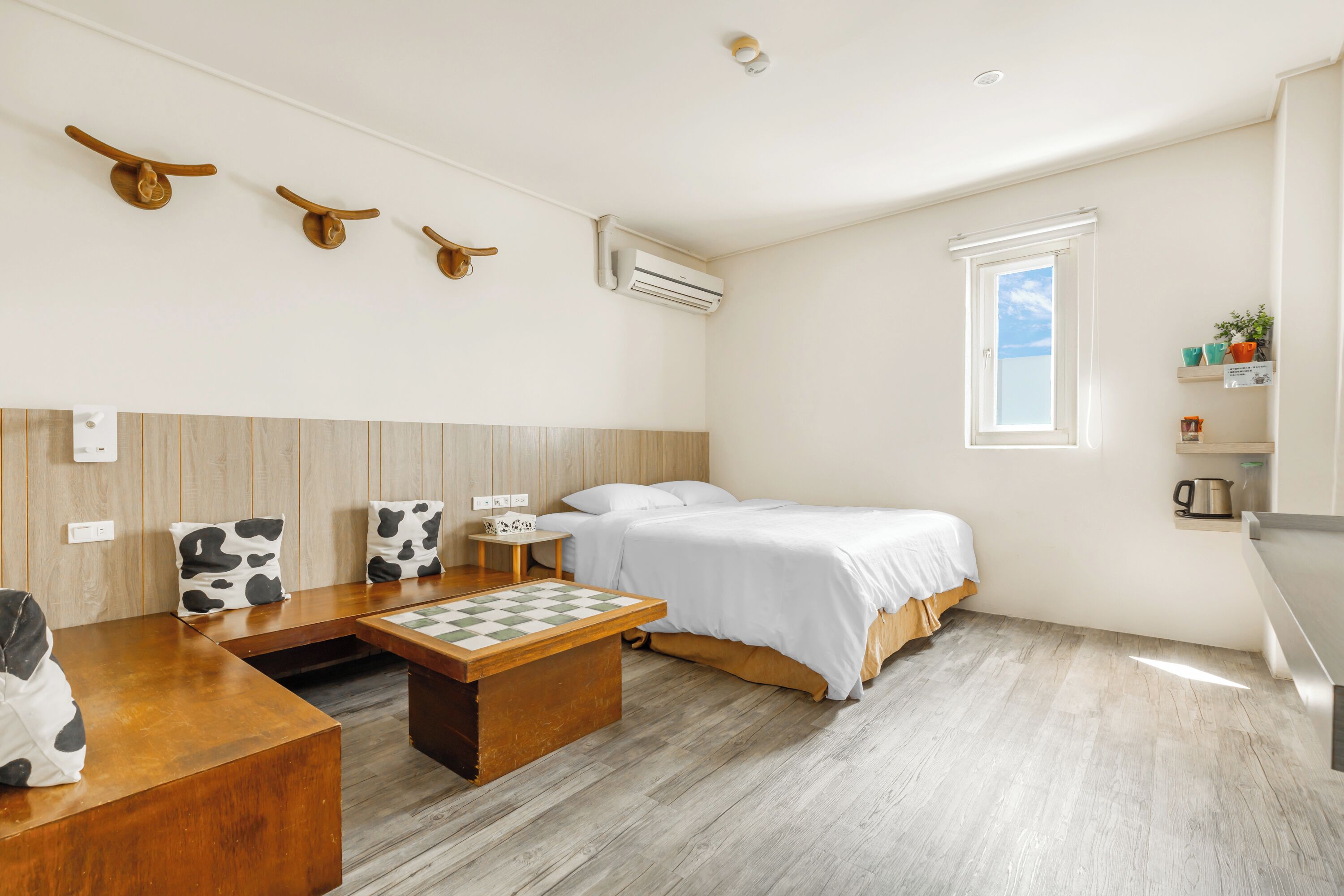 A room with a bed, a wooden bench with a chessboard, a small table, and a window.