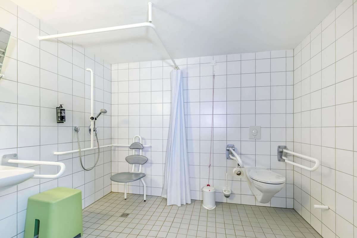 Un baño moderno con paredes de azulejos blancos, una ducha con cabezal de ducha de mano, un inodoro con barra de apoyo y un taburete verde.