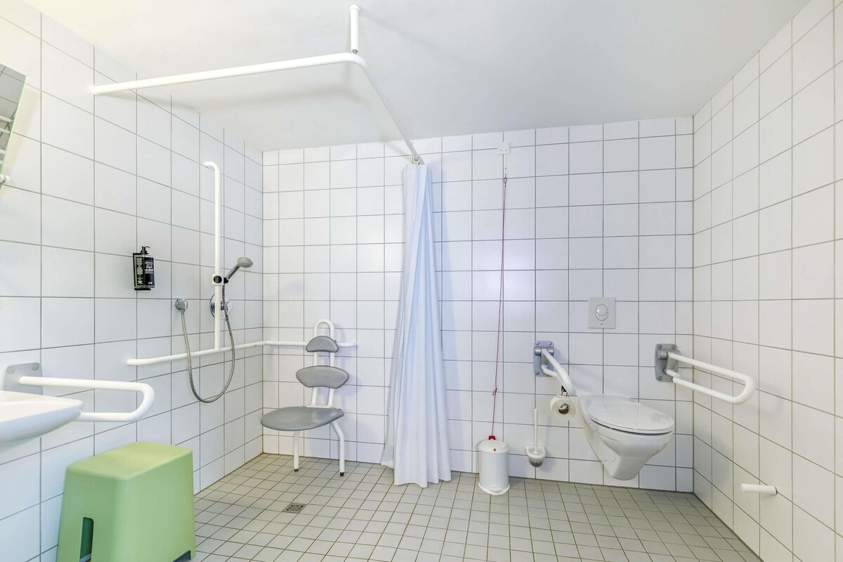 Un baño moderno con paredes de azulejos blancos, una ducha con cabezal de ducha de mano, un inodoro con barra de apoyo y un taburete verde.