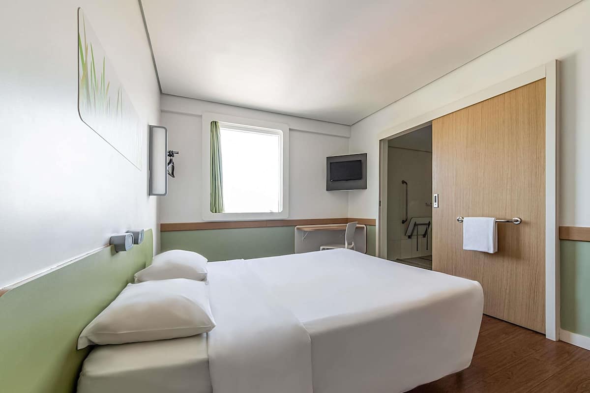 Una habitación de hotel con una cama, televisión, ventana con cortinas, puerta y zona de ducha.