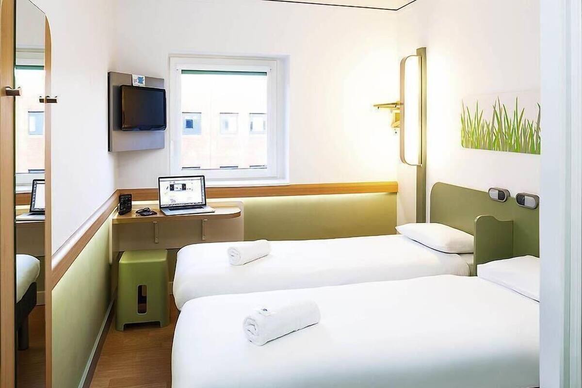 Una habitación de hotel con dos camas, un escritorio con una computadora portátil, un televisor y una ventana.