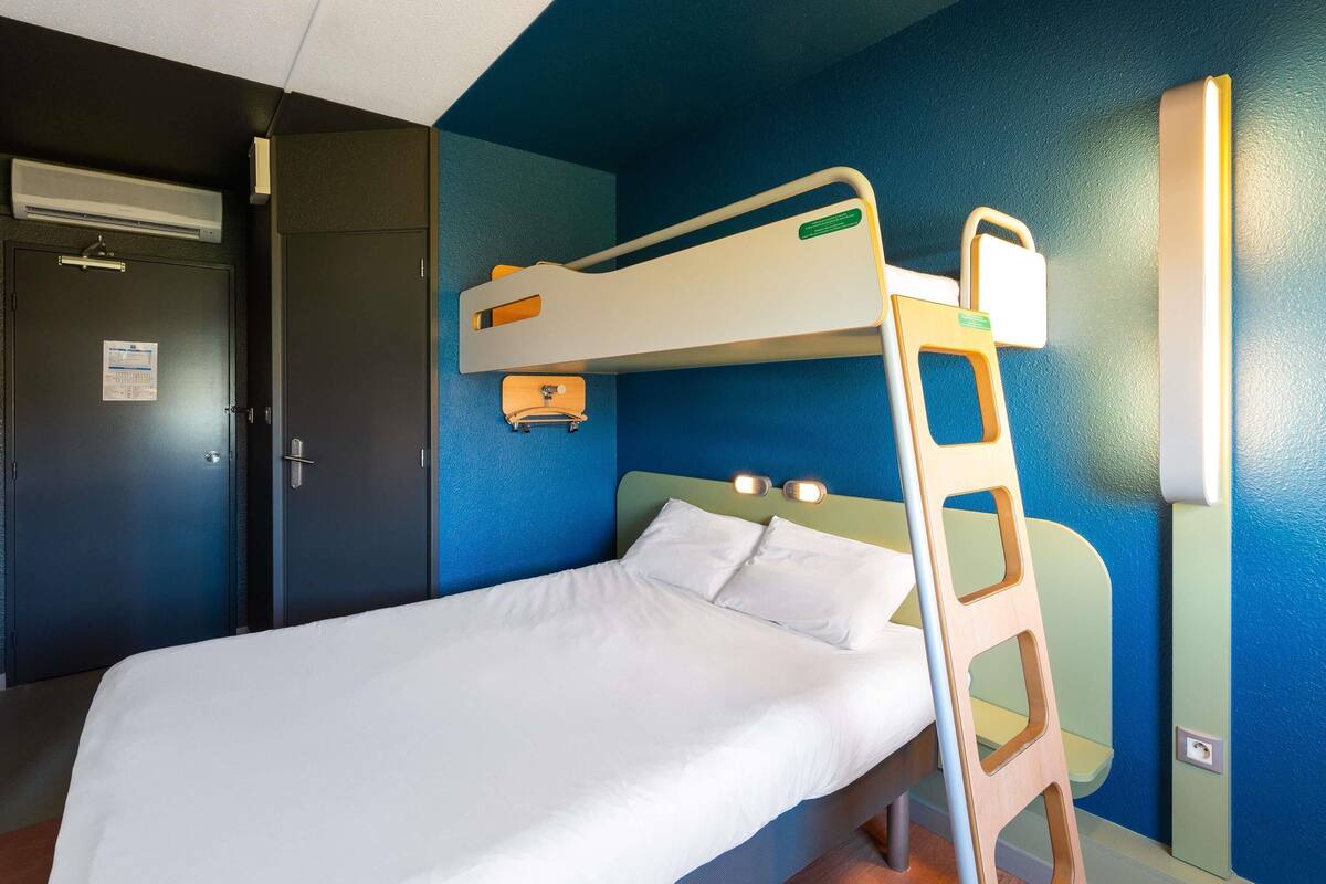 Una cama individual en un dormitorio con pared azul, una puerta con un letrero y un estante pequeño.