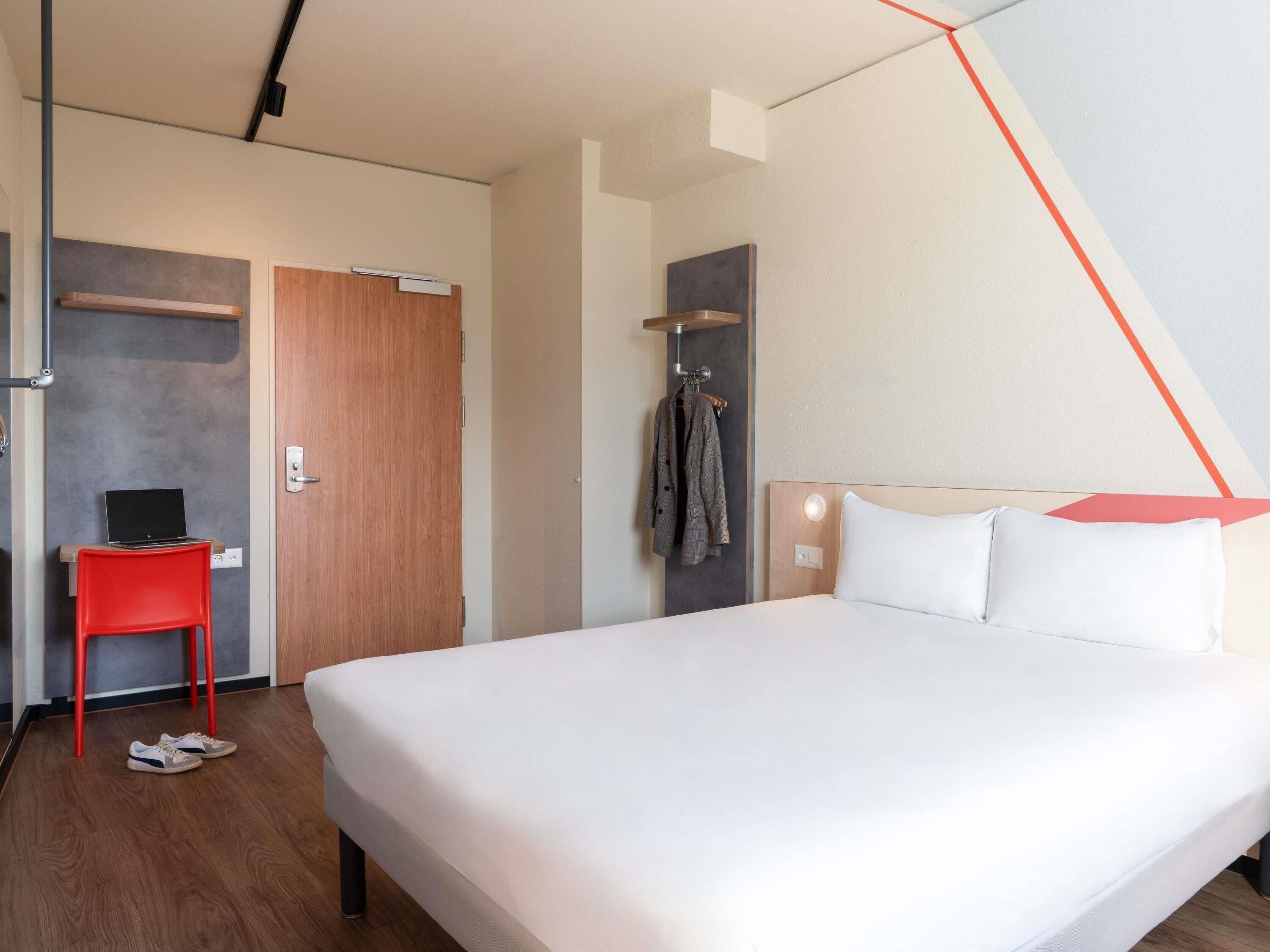 Habitación de hotel con una cama grande, un escritorio con una computadora portátil, una silla roja y un perchero.