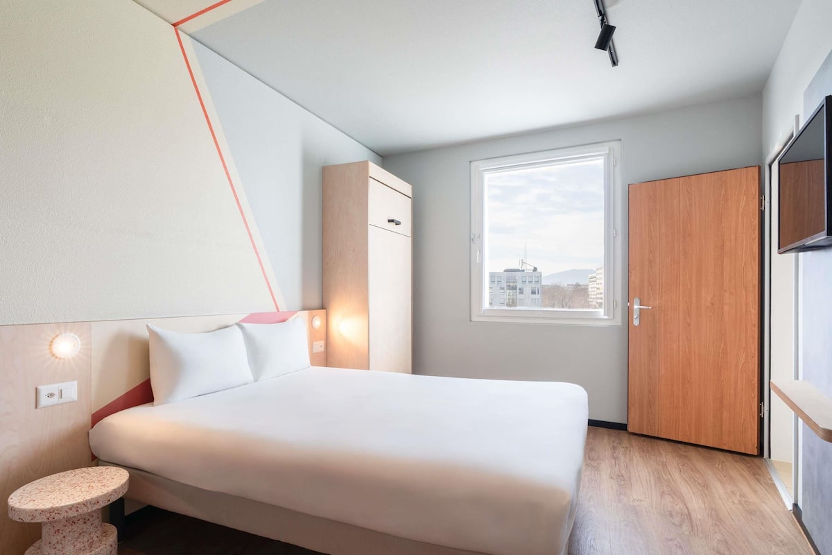 Ein modernes Hotelzimmer mit einem Bett, einem kleinen runden Tisch, einem Schrank und einem Fenster mit Blick auf die Stadt.