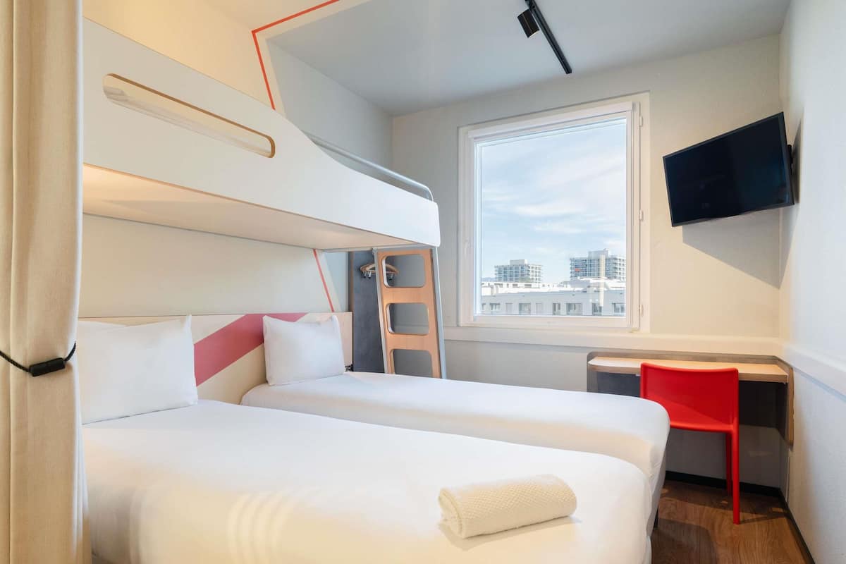 Ein Hotelzimmer mit zwei Betten, einem Schreibtisch, einem Sessel, einem Fernseher und einem Fenster mit Blick auf die Stadt.