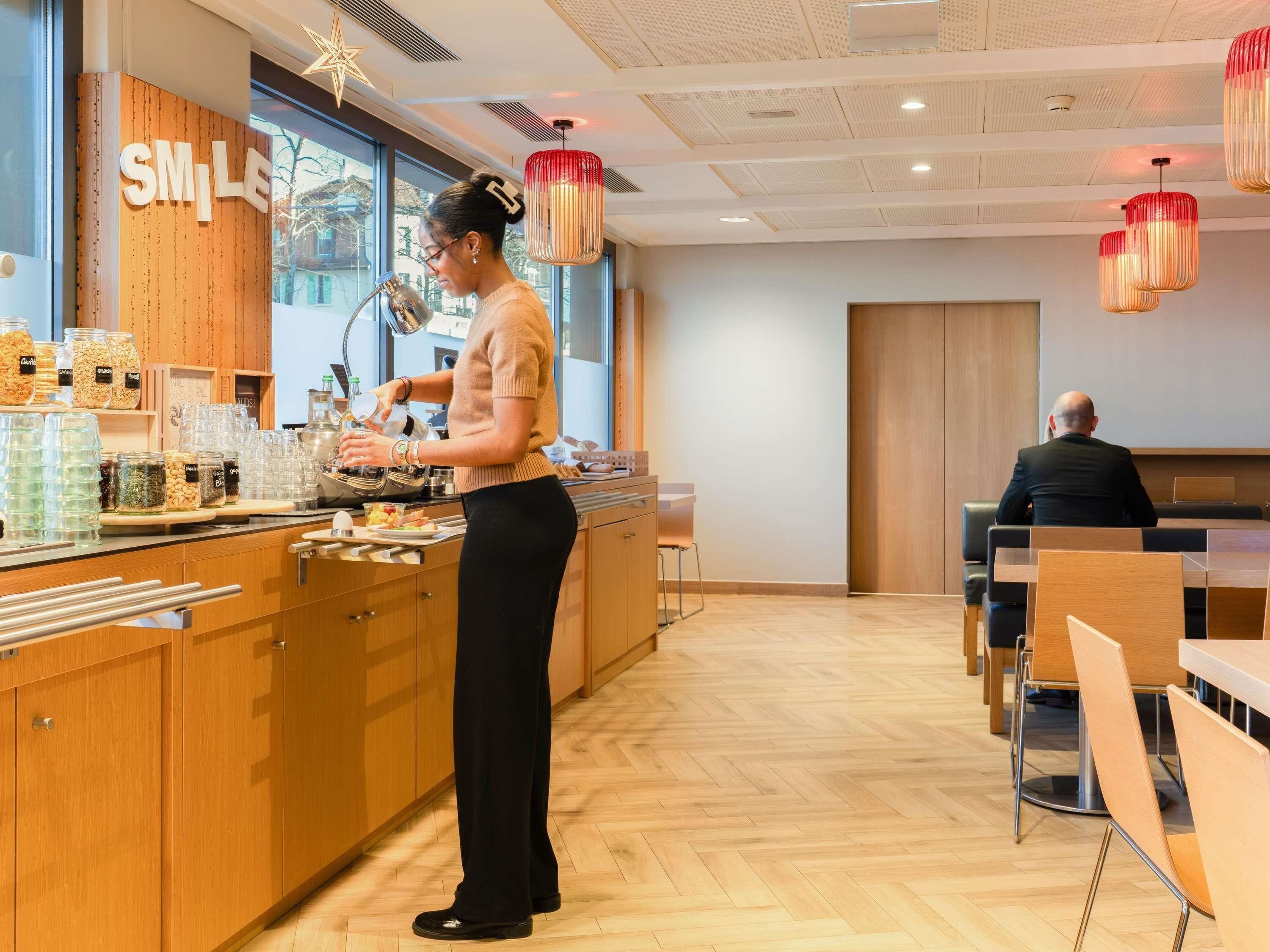 Een café met een toonbank waarop verschillende etenswaren staan uitgestald, en een persoon die eten bereidt. Er is een zitgedeelte met tafels en stoelen.