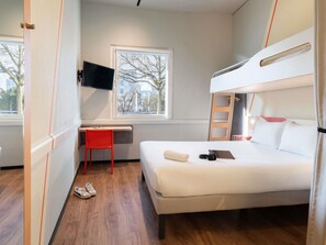 Una habitación de hotel moderna con una cama litera, un escritorio con una silla, un televisor montado en la pared y una ventana con vistas a árboles y edificios.