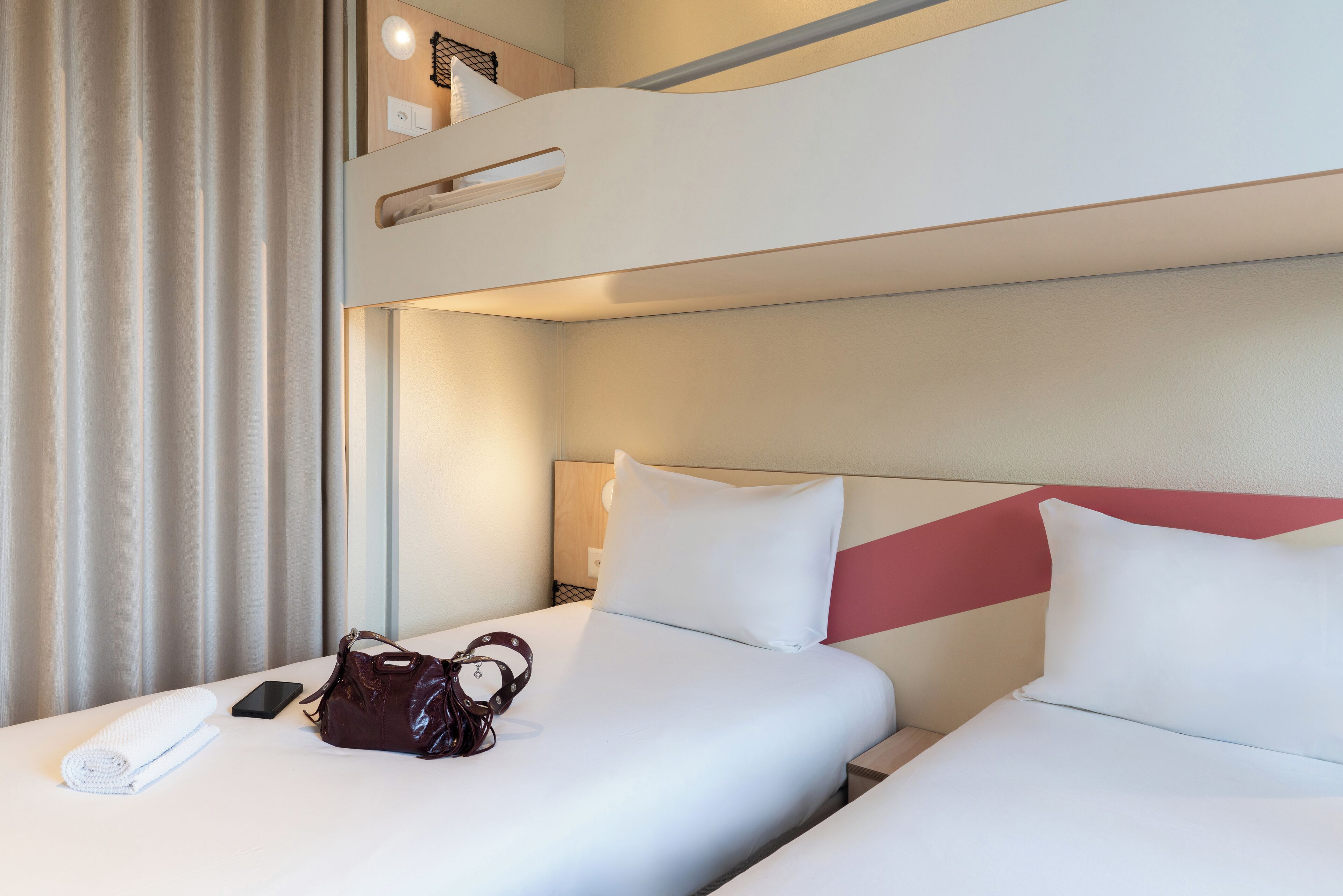 A hotel room with two beds, a red accent wall, and a neatly made bed with a rolled towel and a handbag.