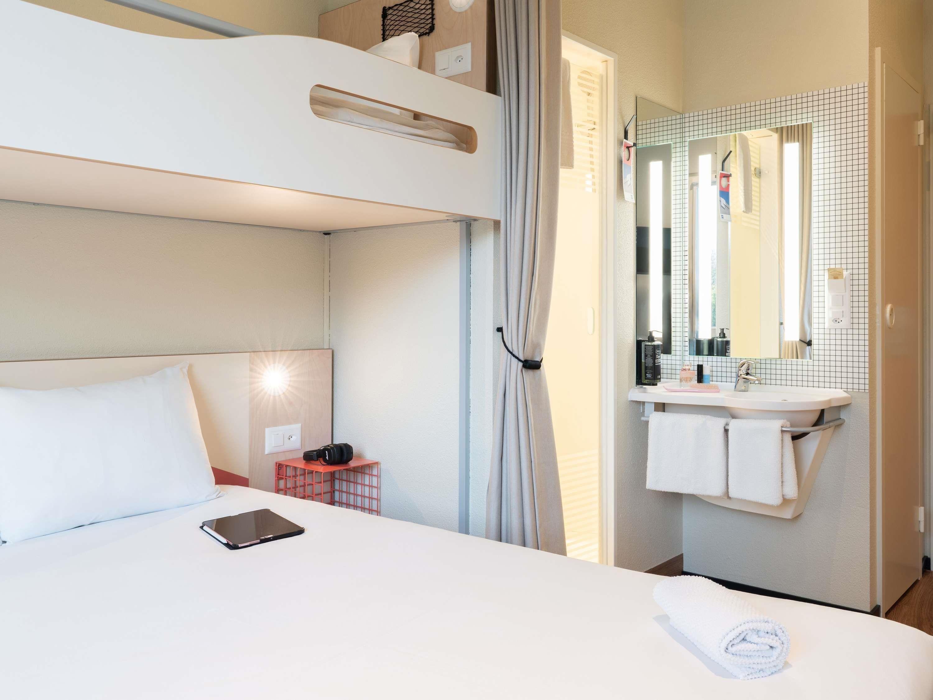Une chambre d’hôtel avec un lit superposé, une table de chevet avec une tablette, une salle de bain avec un lavabo et un miroir, et un porte-serviettes.