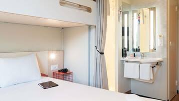 Une chambre d’hôtel avec un lit superposé, une table de chevet avec une tablette, une salle de bain avec un lavabo et un miroir, et un porte-serviettes.