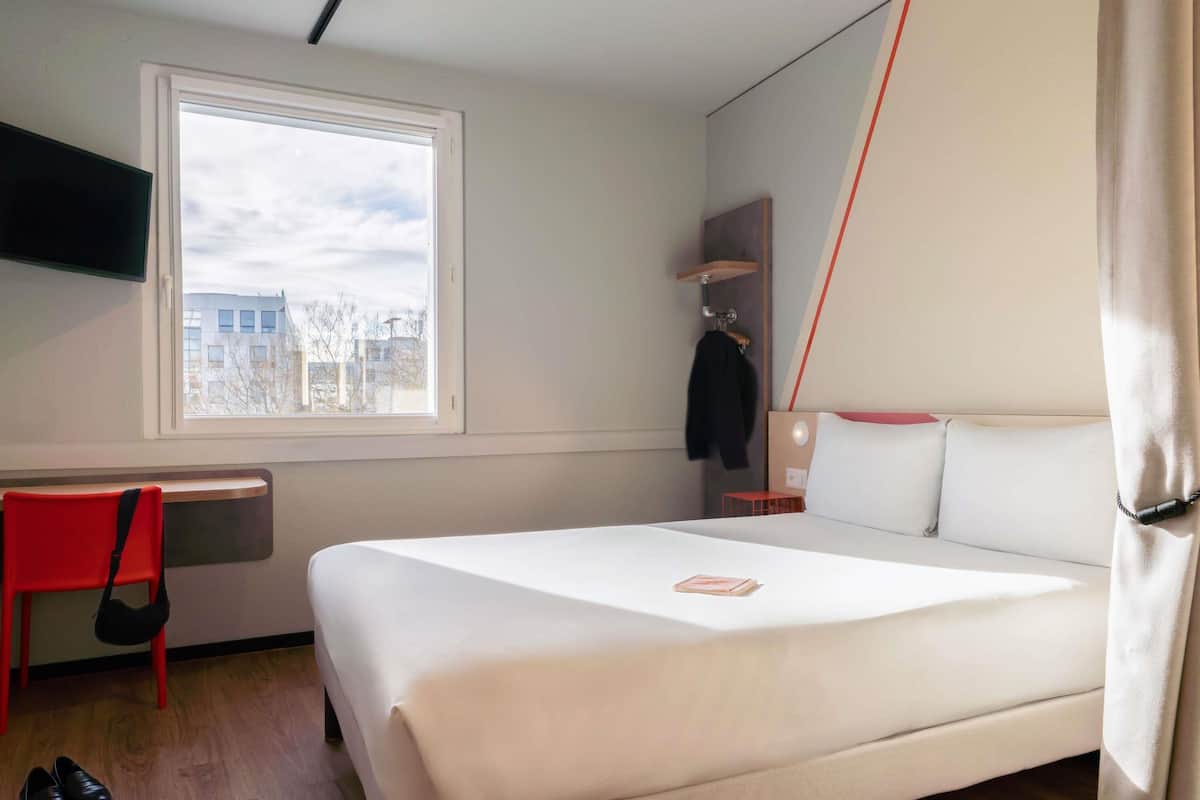 Ein Hotelzimmer mit Bett, Schreibtisch, Stuhl, Fenster und Blick auf Gebäude.