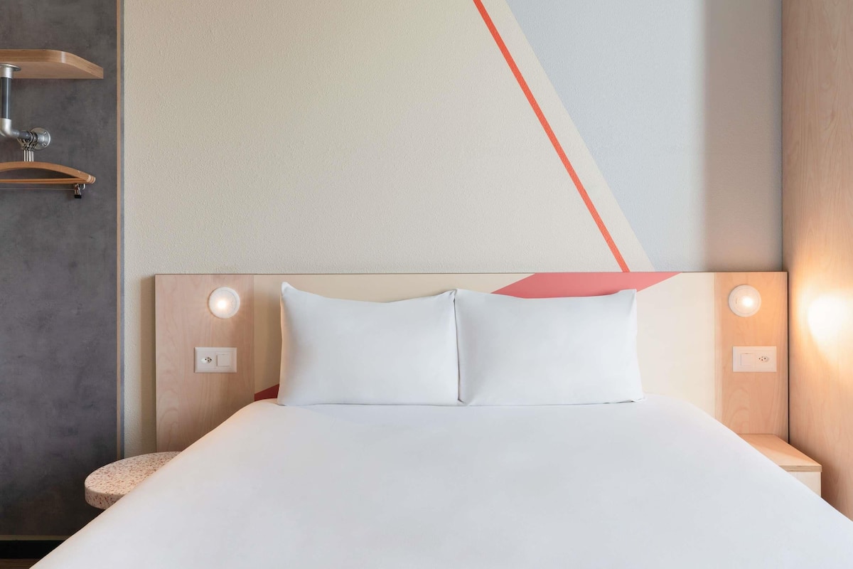 Una habitación de hotel con cama, mesitas de noche, una lámpara de pared y un perchero para toallas.
