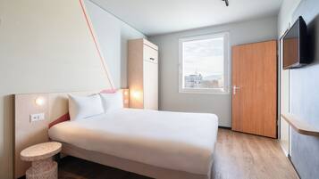 Ein modernes Hotelzimmer mit einem Bett, einem kleinen runden Tisch, einem Kleiderschrank, einem Fernseher und einem Fenster mit Blick auf Gebäude.