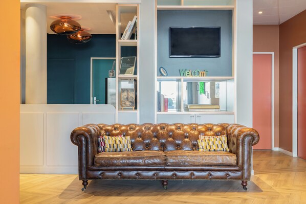 A brown leather sofa with patterned cushions, a television mounted on the wall, and a bookshelf with various items. - ibis budget Geneve Petit Lancy (Lancy)