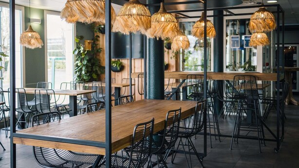 Un interior de restaurante moderno con mesas de madera, sillas de malla metálica negra y lámparas colgantes tejidas.