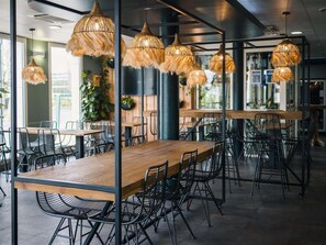 Un intérieur de restaurant moderne avec des tables en bois, des chaises en treillis métallique noir et des suspensions lumineuses en fibres tressées.