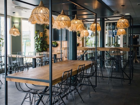 A modern interior with wooden tables and black wire mesh chairs, hanging woven light fixtures, and large windows allowing natural light.. ibis budget Bordeaux Aeroport