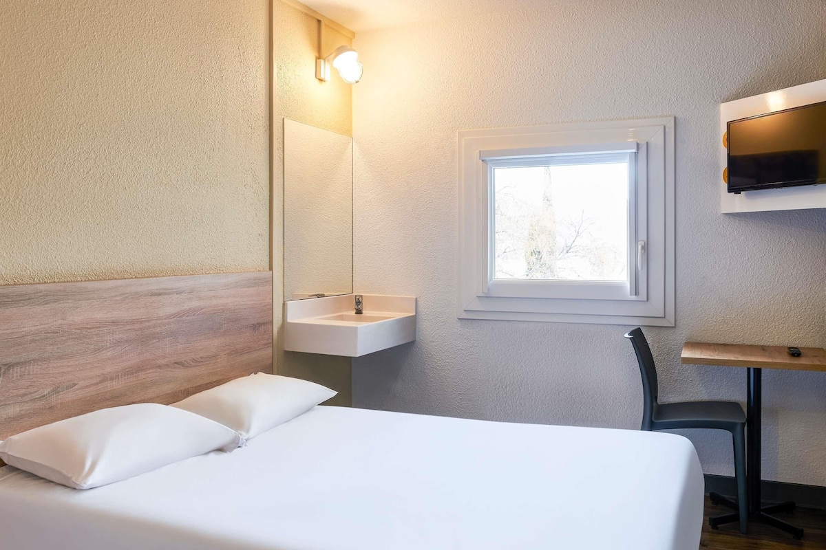 A bedroom with a bed, a bedside table, a mirror, a window, and a sink.