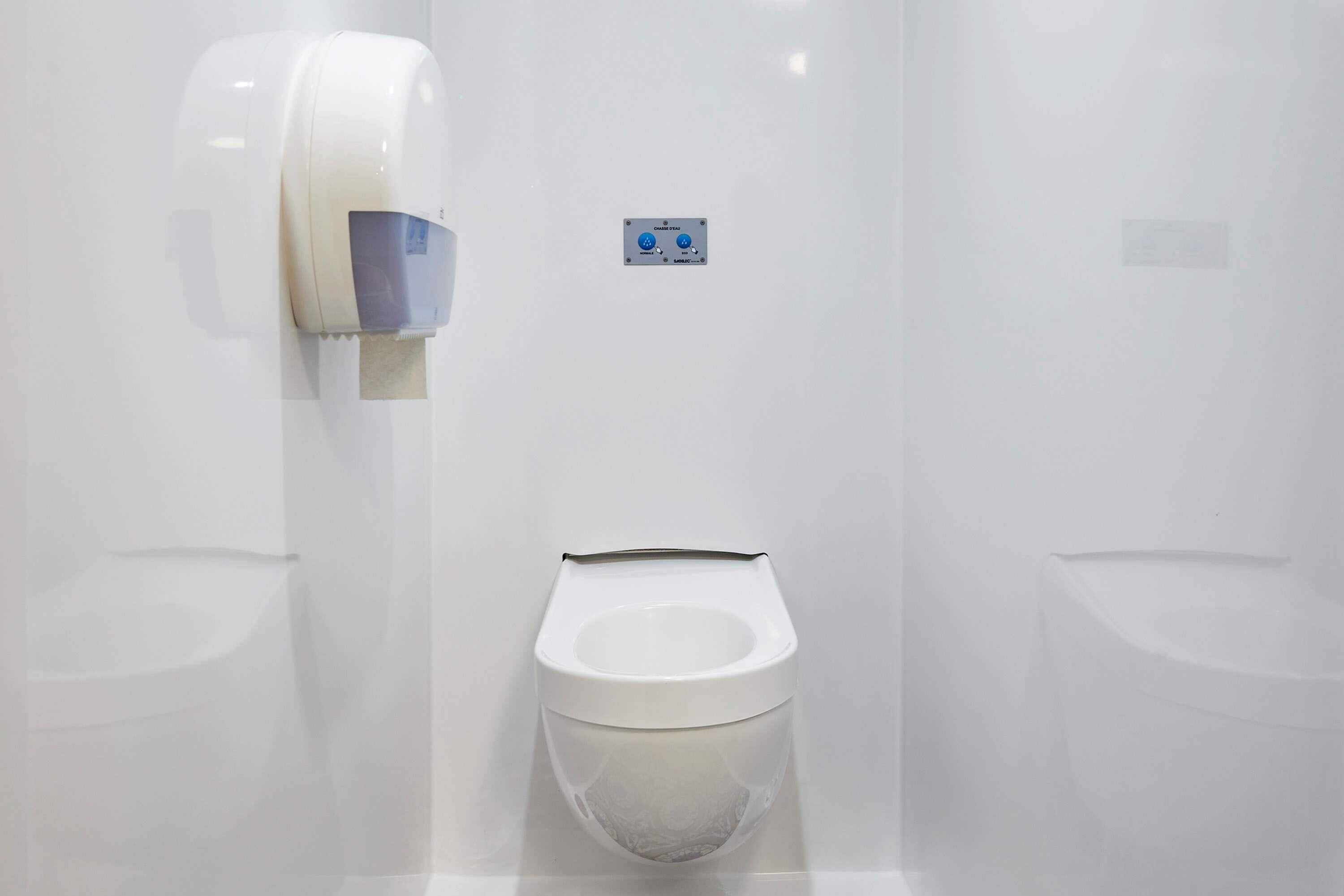 Un baño moderno y blanco con dispensador de papel higiénico en la pared y botón de descarga.