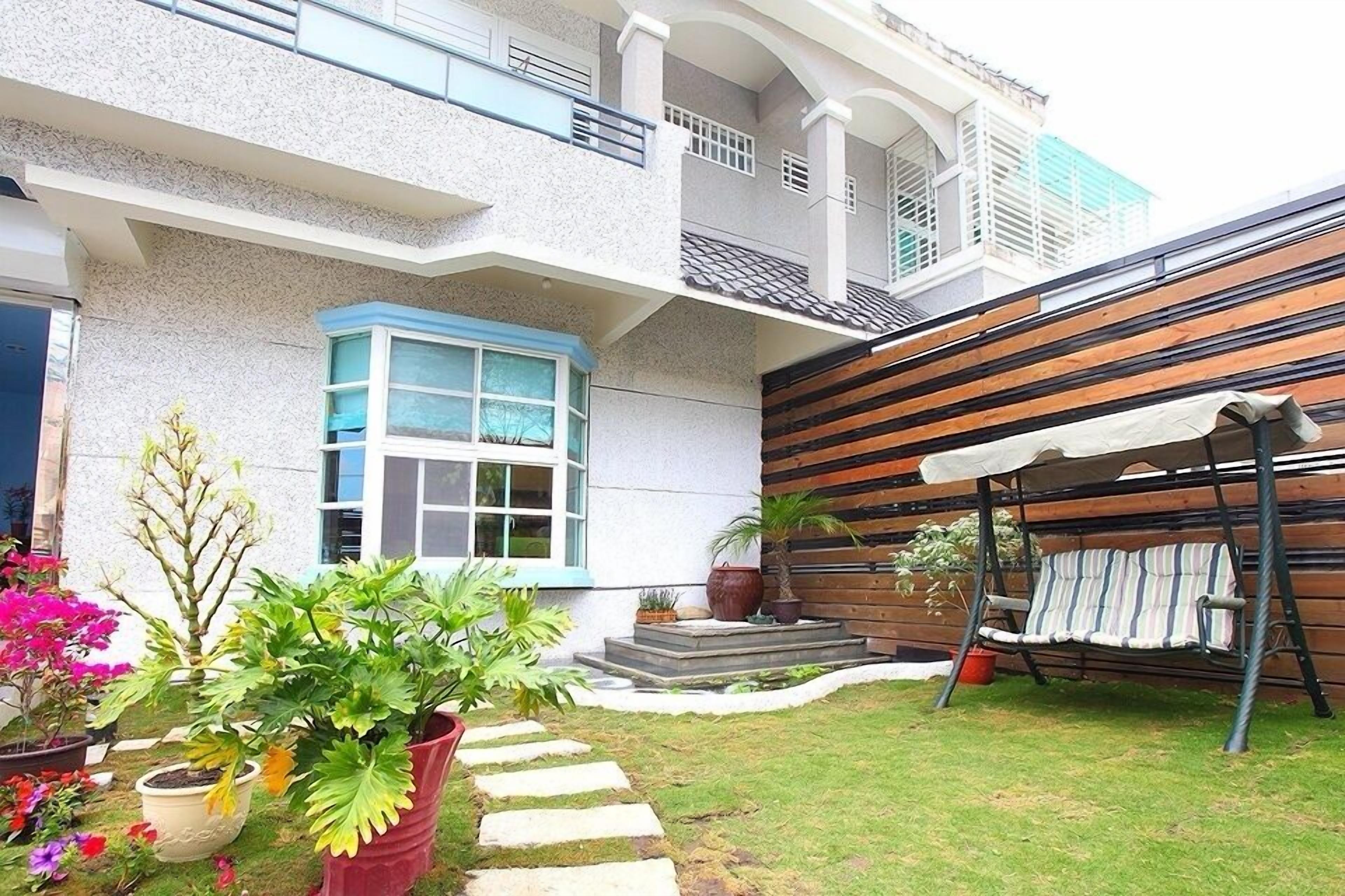 A modern two-story house with a balcony, a wooden fence, and a garden with potted plants and a swing.