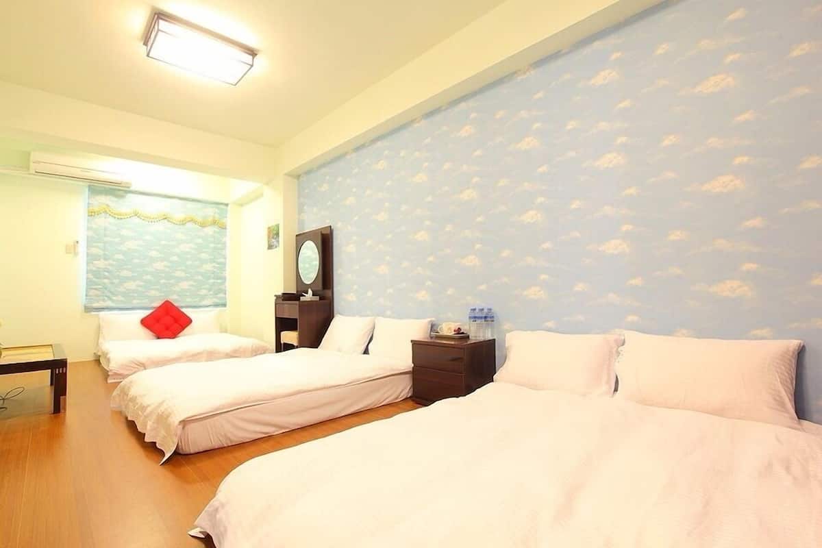 Habitación de hotel con dos camas, suelo de madera, pared con diseño de nubes, ventana con vista al mar y una mesita con un cojín rojo.