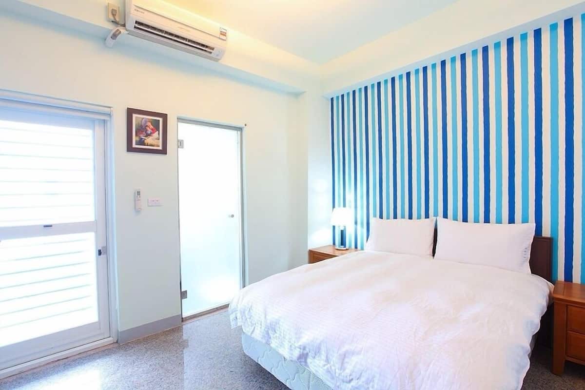 Una habitación de hotel con una cama, una mesita de noche, un aire acondicionado de pared y una puerta con ventanal, además de un muro a rayas.