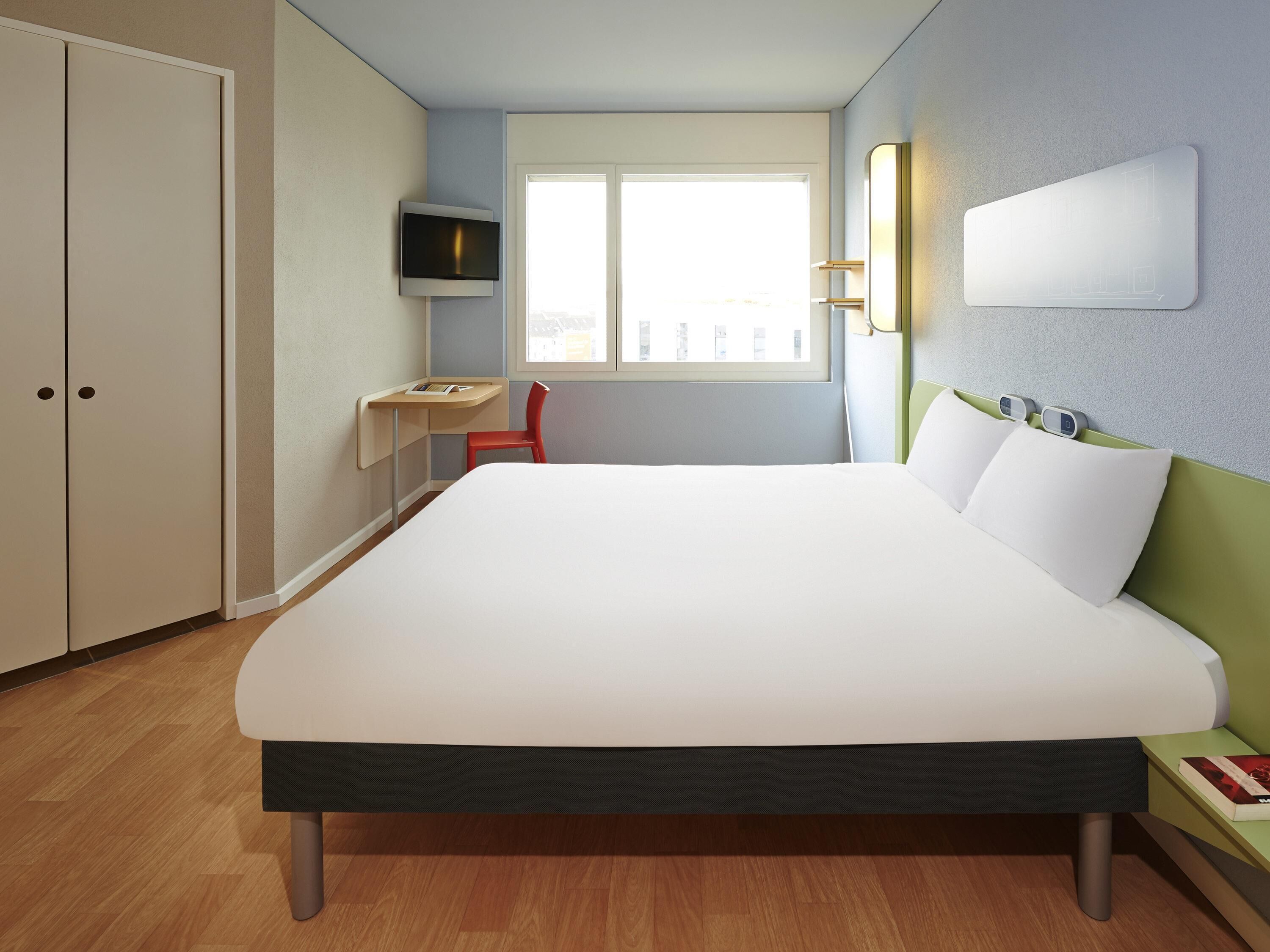 A hotel room with a bed, a desk, a chair, a TV, and a window.