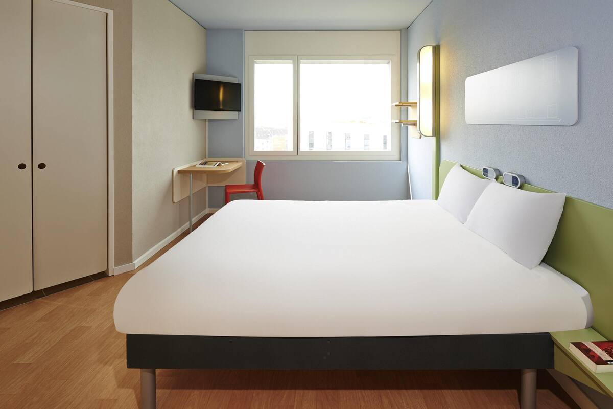 A hotel room with a bed, a desk, a chair, a TV, and a window.