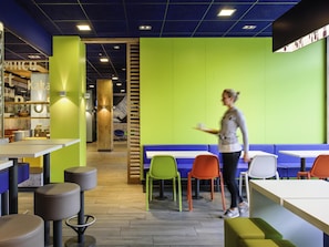 A modern cafe with wooden flooring, green walls, and a variety of seating options including tables and chairs. - ibis budget Basel City (Basel)