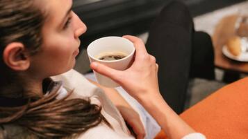 Eine Person hält eine Tasse, vermutlich mit einem Getränk, und sitzt in einem Zimmer mit einem Fenster und einem Tisch mit Teller und Glas.