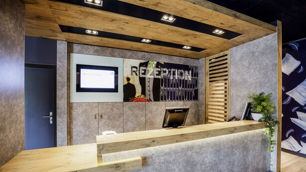 A reception desk with a wooden counter, a digital screen, and a wall-mounted display. - ibis budget Basel City (Basel)