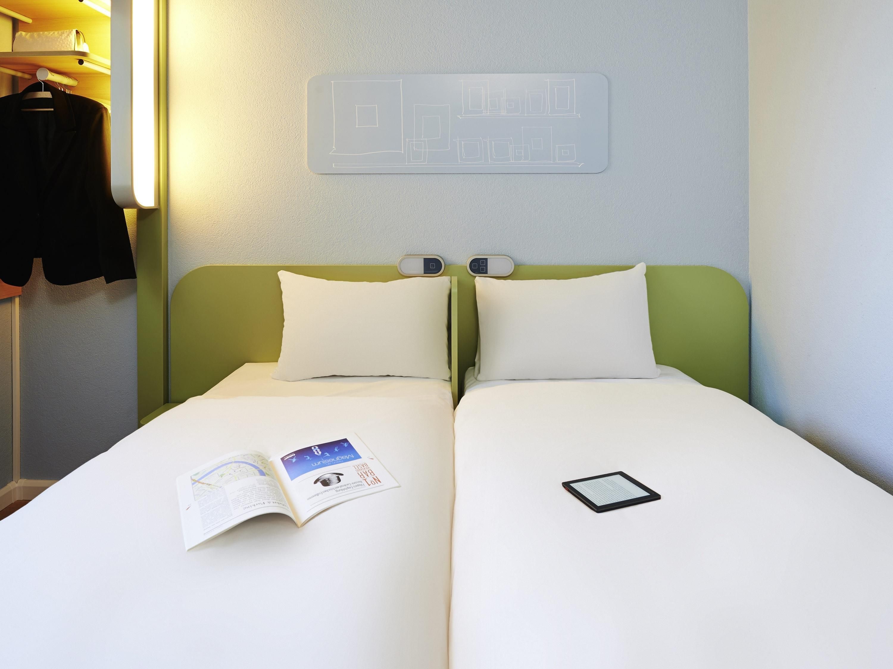 A double bed with white bedding, a bedside table with a book and a tablet, and a wall-mounted light fixture.