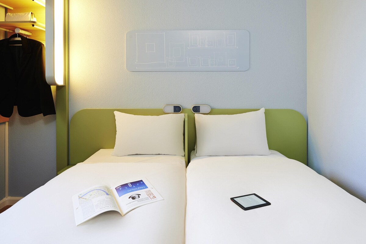 A double bed with white bedding, a bedside table with a book and a tablet, and a wall-mounted light fixture.