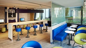 A modern bar area with a wooden counter, multiple stools, and a seating area with round tables and colorful chairs. - ibis budget Basel City (Basel)