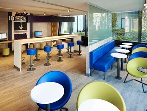 A modern bar area with a wooden counter, multiple stools, and a seating area with round tables and colorful chairs. - ibis budget Basel City (Basel)