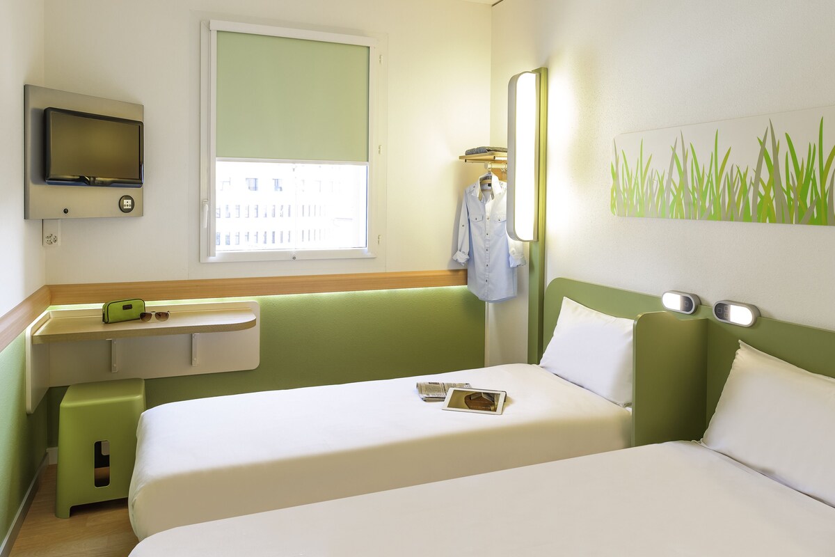 Une chambre d’hôtel comprenant un lit, un téléviseur fixé au mur, une fenêtre avec des stores, une table de chevet avec un téléphone et des lunettes, et une œuvre d’art décorative au mur.
