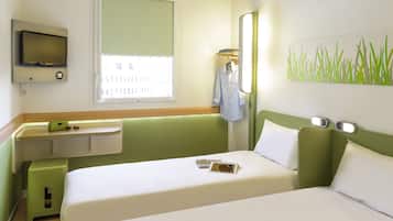 A hotel room with a bed, a TV mounted on the wall, a window with blinds, a bedside table with a phone and glasses, and a wall art piece.
