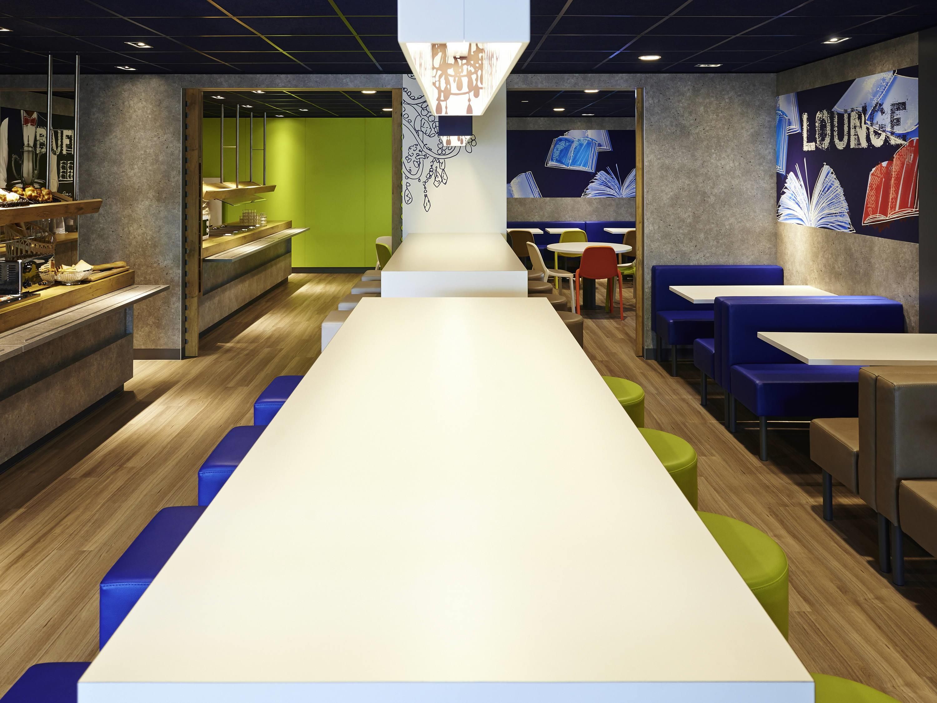 A modern lounge area with a long white table, blue and green seating, and a wall with 'LOUNGE' and book illustrations.