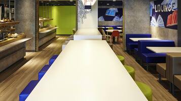 Un moderno espacio de lounge con una larga mesa blanca, asientos azules y verdes, y una pared con la palabra 'LOUNGE' e ilustraciones de libros.