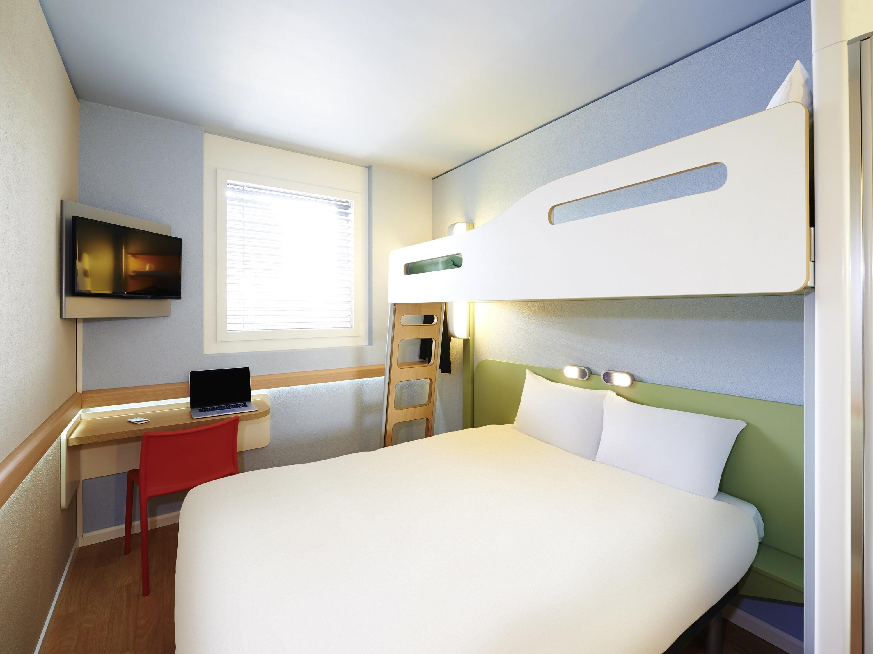A hotel room with a bed, a desk with a laptop, a TV mounted on the wall, and a window with blinds.