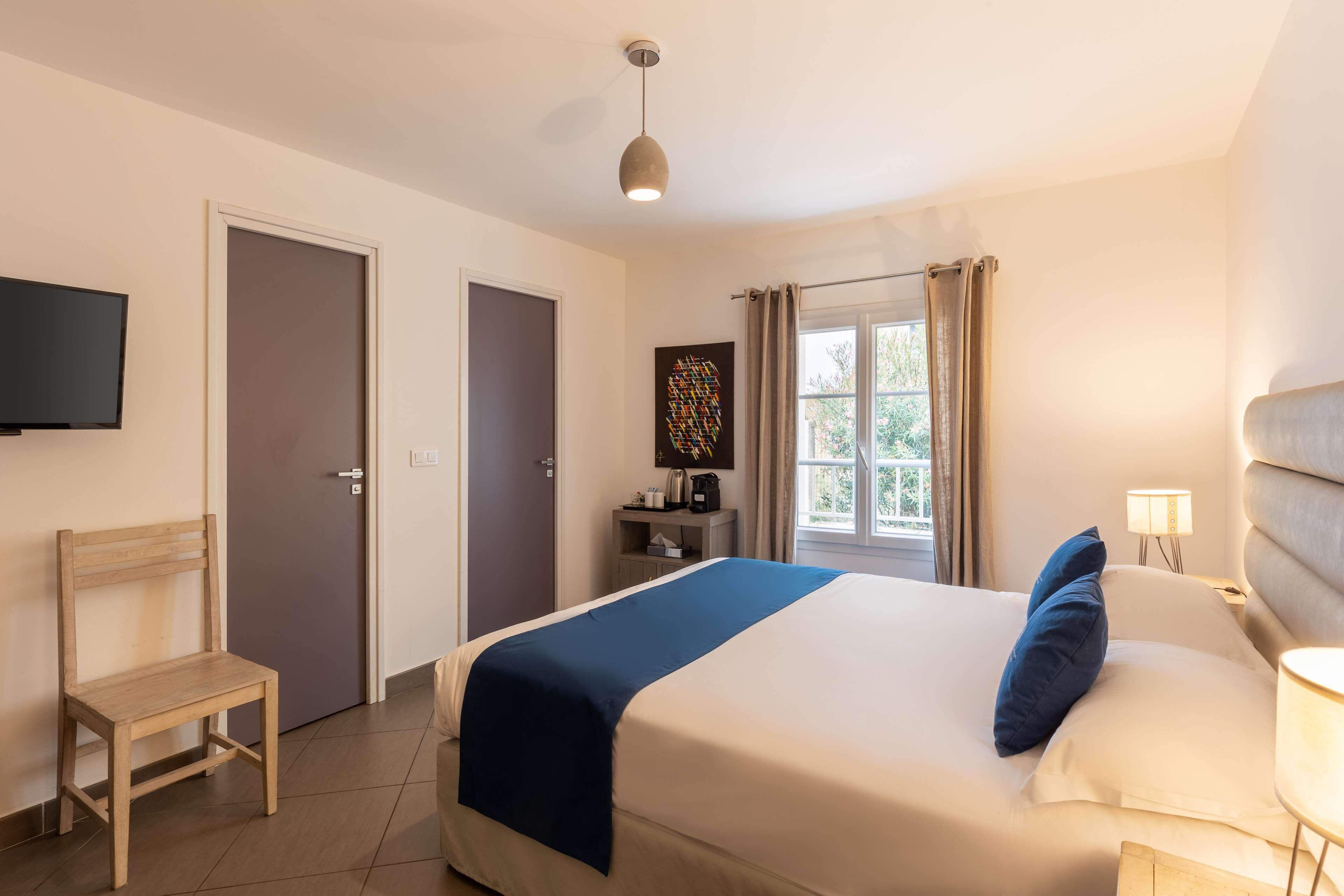 Une chambre d’hôtel avec un lit, une chaise, un bureau avec une lampe, une télévision et une fenêtre avec des rideaux.