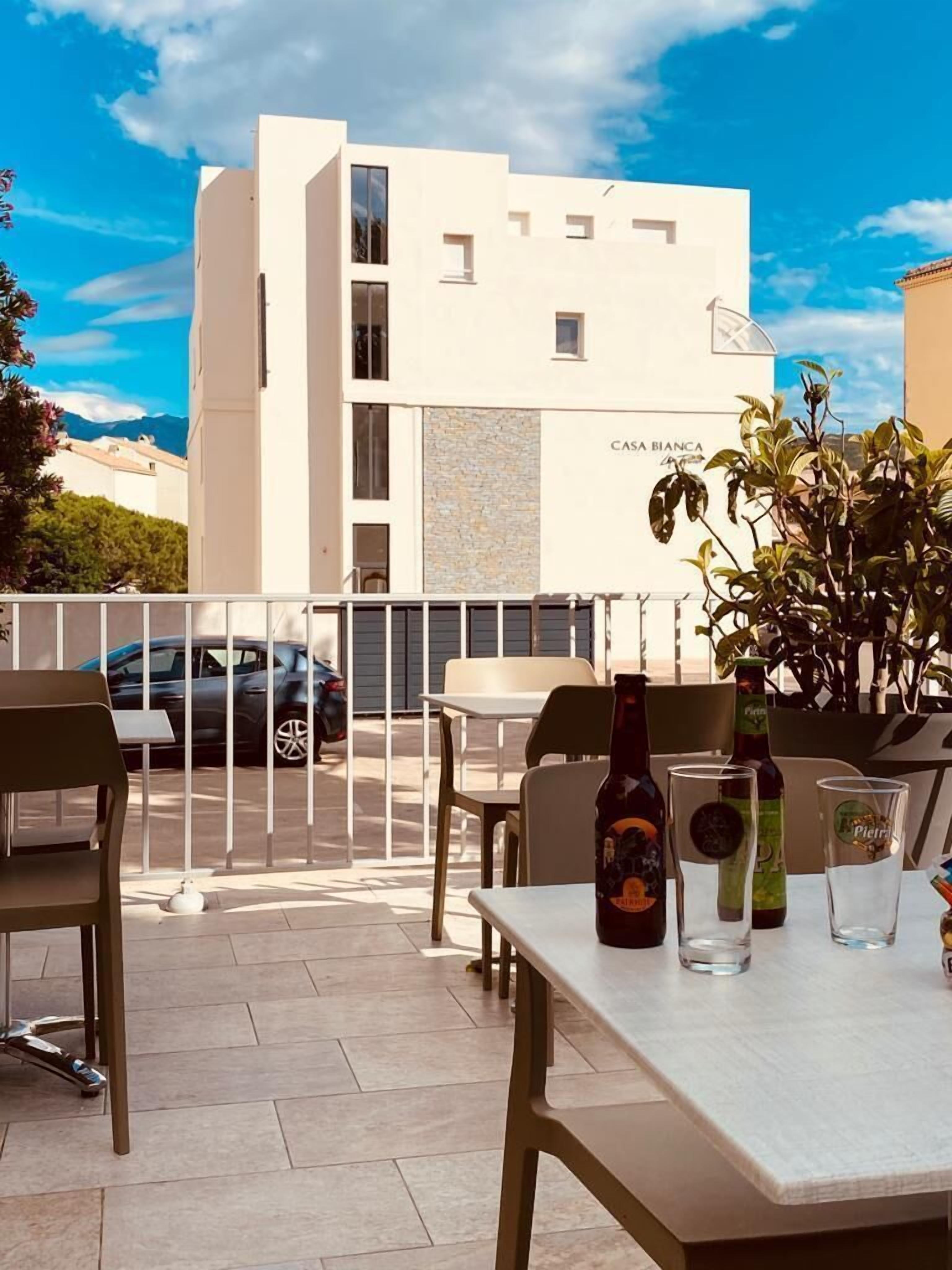 Une terrasse avec des sièges en plein air, une table avec des bouteilles et des verres, et un bâtiment portant l’inscription « CASA BIANCA » en arrière-plan.