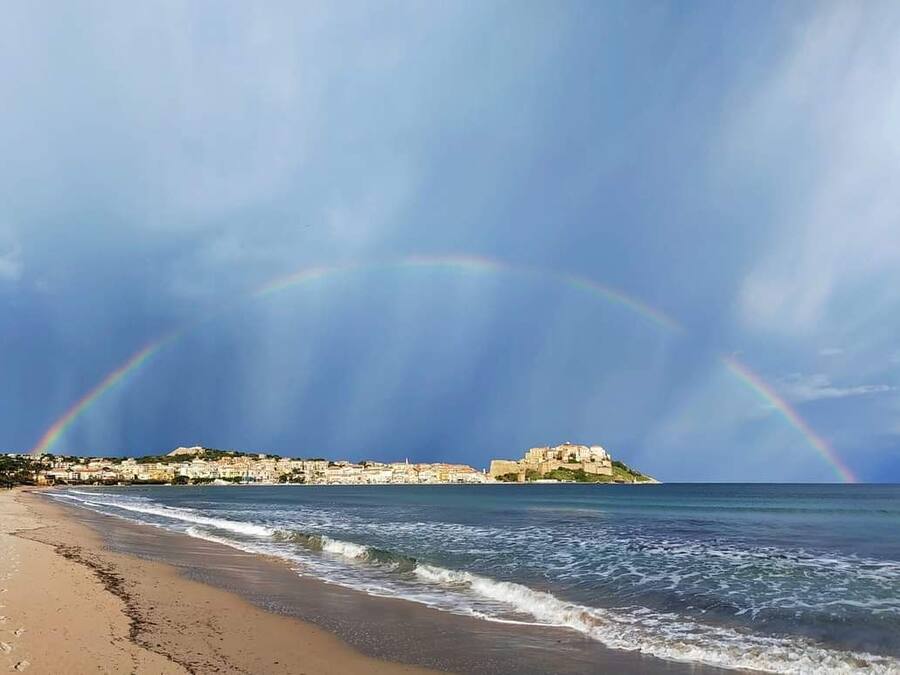A beach with waves, a rainbow, and a building on a hill.