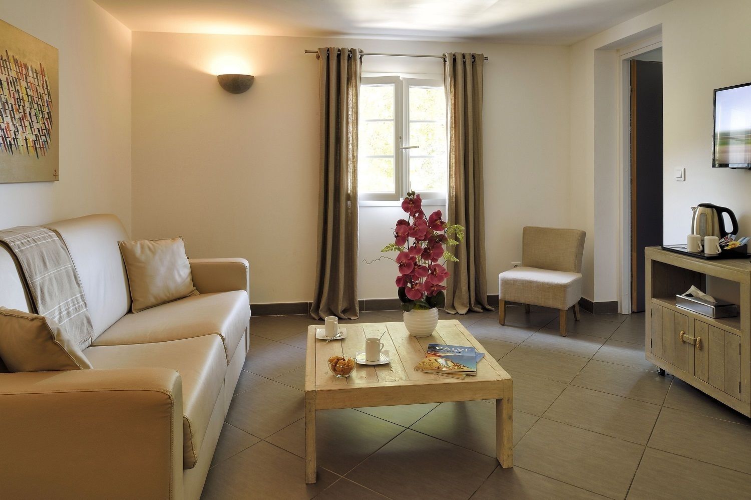 Un salon avec un canapé beige, une table basse en bois sur laquelle se trouvent un livre et une tasse, un fauteuil, un meuble contenant une bouilloire et une fenêtre avec des rideaux.