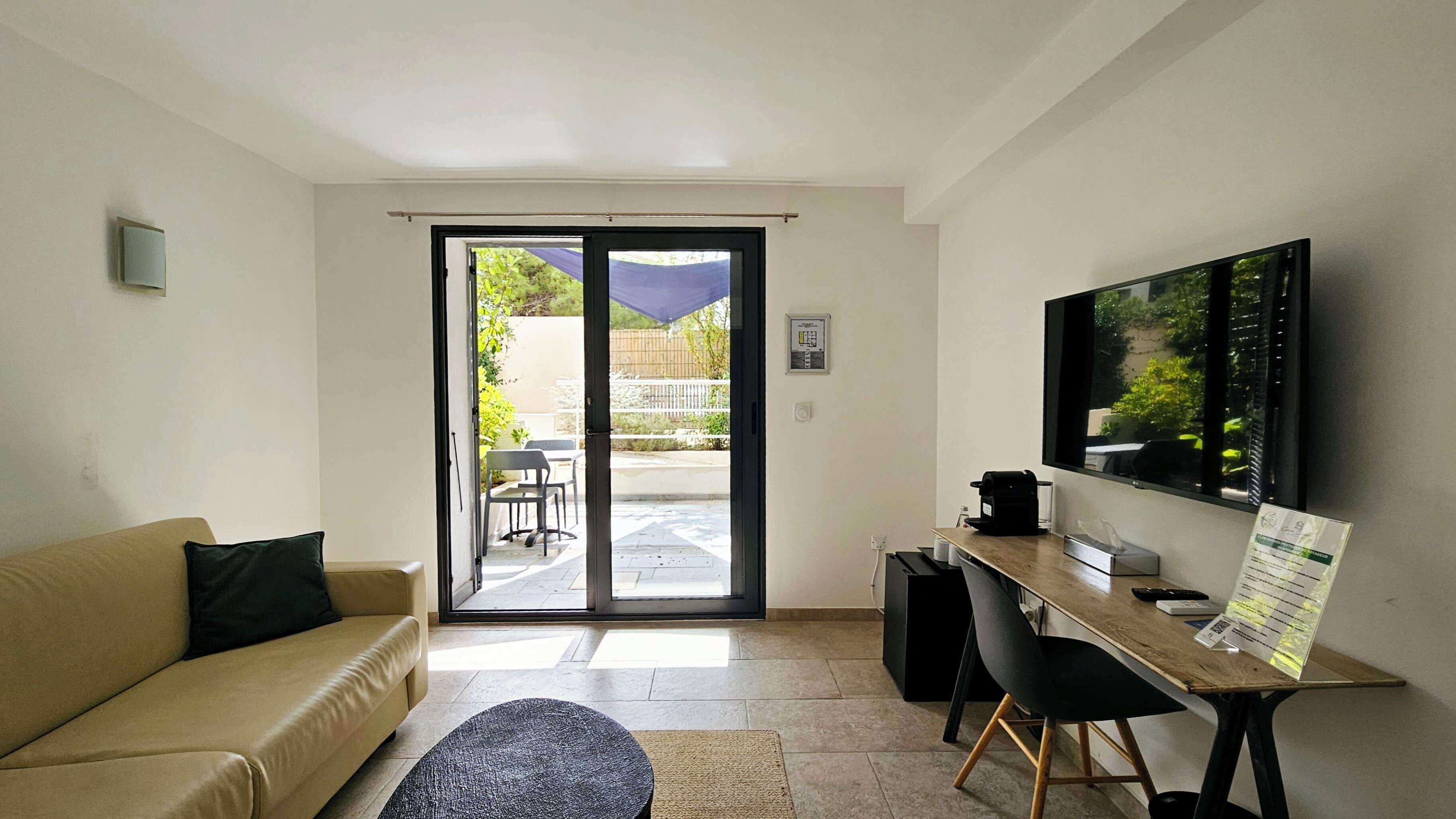 Una sala de estar moderna con un sofá, una mesa de centro, un escritorio con una silla, un televisor de pantalla plana y una puerta corrediza de vidrio que da a un patio al aire libre.