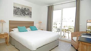 Quarto de hotel com uma cama grande, duas mesas de cabeceira com luminárias, uma cadeira de vime e uma varanda com mesa e cadeiras.