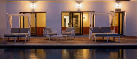 Un área junto a la piscina con cuatro sillones de descanso y una mesa, una pérgola sobre una de las sillas, y un edificio con puerta de vidrio y una lámpara.