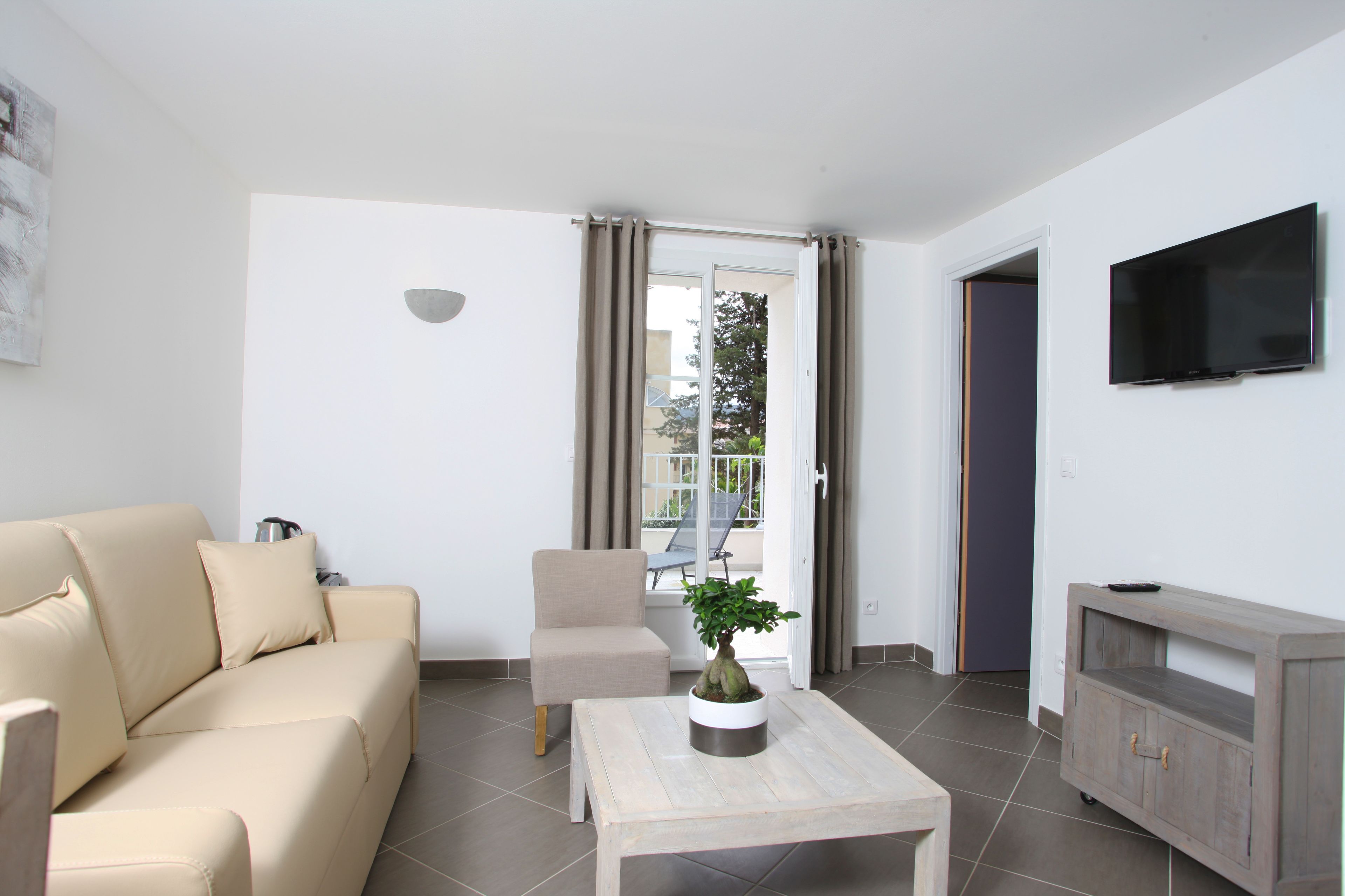 Un salon avec un canapé beige, une petite table sur laquelle est posée une plante, un téléviseur fixé au mur et une porte coulissante donnant sur l’extérieur.
